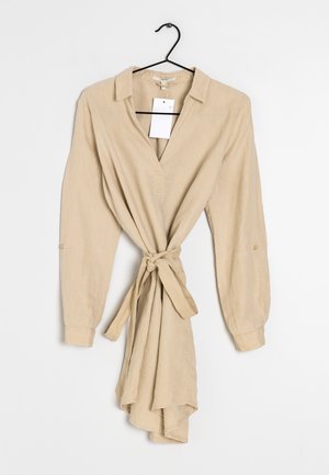 Robe beige à manches longues avec col et ceinture à nouer, suspendue à un cintre noir sur fond blanc.