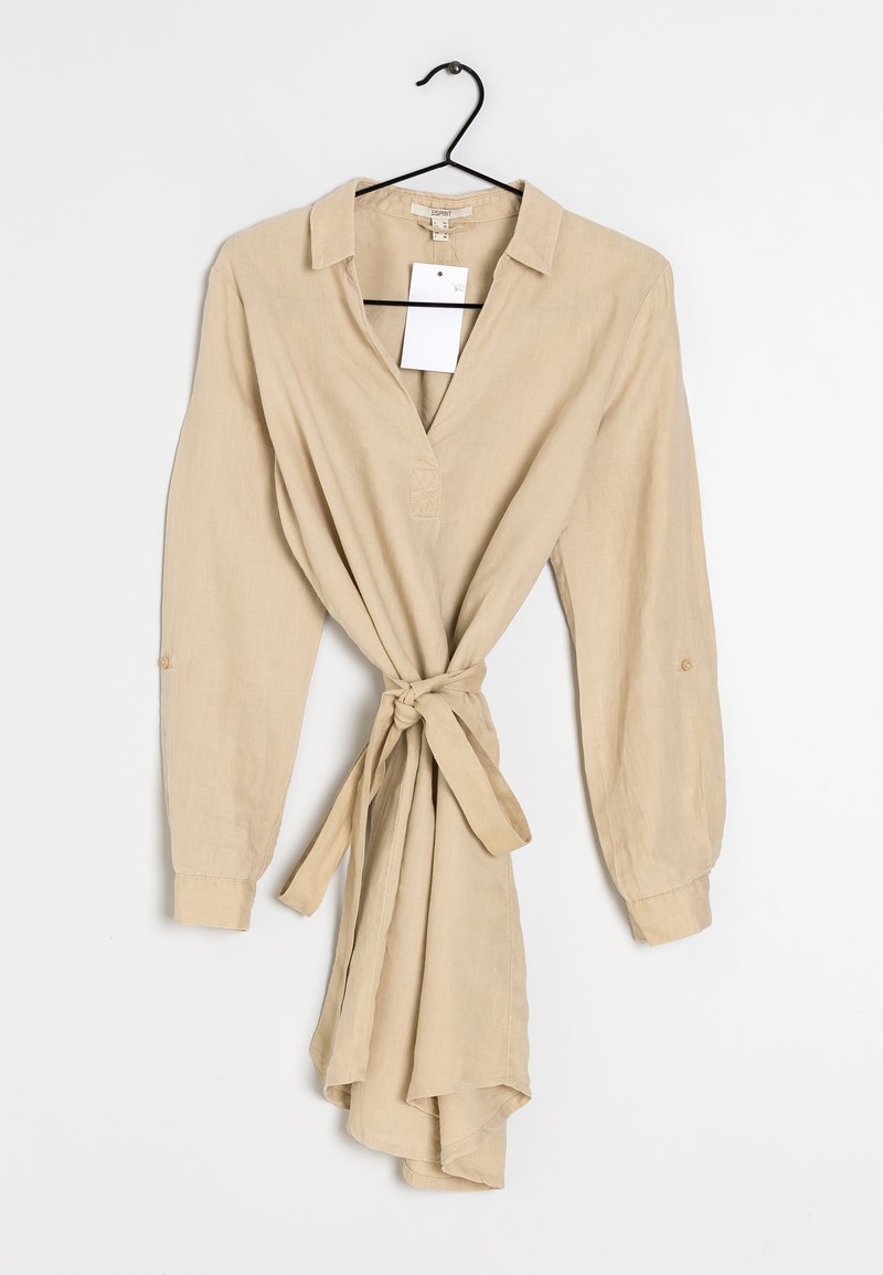 Robe beige à manches longues avec col et ceinture à nouer, suspendue à un cintre noir sur fond blanc.