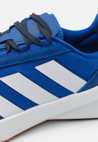 Tenis de tecido azul com design de três riscas brancas, apresentando um upper texturizado, atacadores cinzas e uma sola de borracha branca.