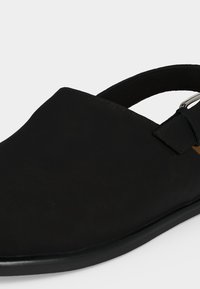 Μαύρο παπούτσι χωρίς κορδόνια με κλειστή μύτη, ανοιχτή φτέρνα και ρυθμιζόμενο λουράκι στον αστράγαλο με ασημένια αγκράφα.