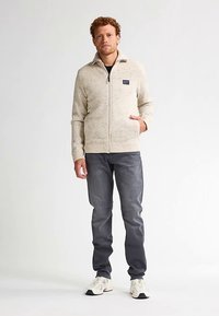 Pulover bej deschis cu fermoar, tricotat texturat, cu o etichetă pe pieptul stâng, asortat cu blugi gri închis și adidași albi. Croială casual.