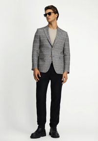 Grijze geblokte blazer met gelapte revers, bruine knopen, gecombineerd met een lichtgrijze trui en zwarte broek, afgewerkt met zwarte laarzen.