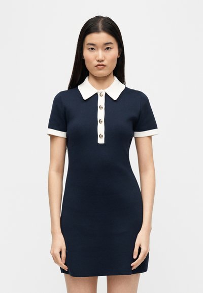Woman with long dark hair wearing a navy blue short-sleeve dress with a white collar, white button placket, and white sleeve trim.