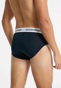 Uomo che indossa boxer navy blu con cintura bianca etichettata "GUESS JEANS", visto da dietro, su uno sfondo chiaro e semplice.