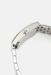 Reloj de metal plateado con una caja rectangular, perfil plano y una correa de eslabones. Presenta una corona redonda y una superficie pulida.