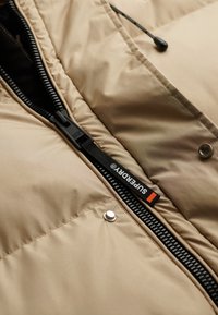 Beige puffjakke med quiltet tekstur, sort lynlås, sølvfarvede knapdetaljer og kontrastfarvet logo-mærke. Glat, isoleret stof.
