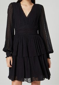 Svart klänning med V-ringning och långa, genomskinliga puffärmar. Har en texturerad tyg med ett lager-på-lager kjol och instoppade detaljer vid midjan.