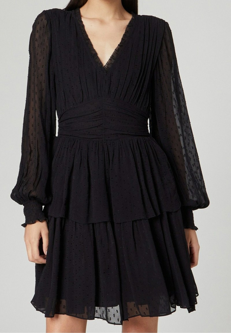 Svart klänning med V-ringning och långa, genomskinliga puffärmar. Har en texturerad tyg med ett lager-på-lager kjol och instoppade detaljer vid midjan.
