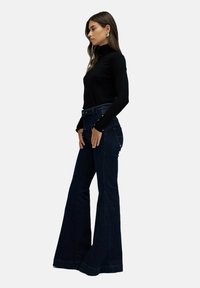 Jeans de campana en un azul oscuro con cintura alta, con botones de latón y detalles de costura, combinados con un top negro ajustado de manga larga.
