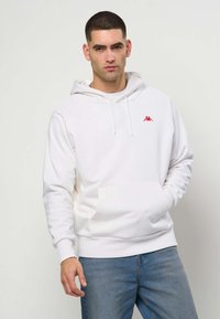 Noor mees lühikeste tumedate juustega, kes kannab valget kapuutsiga pusa punase logoga ja helesiniseid teksaseid, seisab ühe käega taskus.