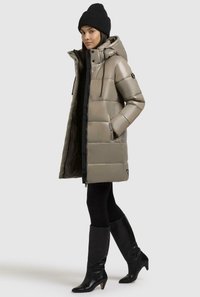 Cappotto imbottito beige con cappuccio, caratterizzato da una finitura lucida, design trapuntato, zip frontale e tasche laterali. Indossato con leggings neri e stivali.