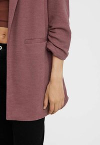 Dlouhý oversize blazer v mauve texturované tkanině s náprsní kapsou, kombinovaný s přiléhavým hnědým topem a černými džíny, na kterém je vidět ruka odpočívající.