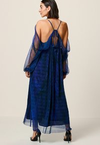 Rochie maxi fluidă, de culoare albastră, cu mâneci transparente, spate descoperit, detaliu cu legătură și un model texturat al materialului; prezintă o siluetă lejeră și lungime până la glezne.