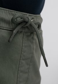 Khaki groene shorts met een trekkoord in de taille, voorzien van een stevige textuur en metalen uiteinden op het trekkoord, gepresenteerd tegen een neutrale achtergrond.