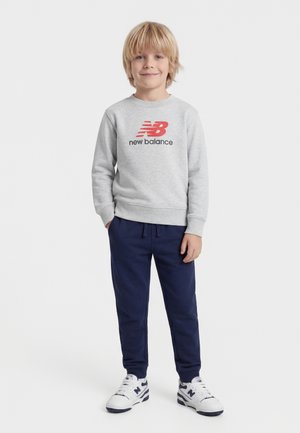 Ragazzo biondo in piedi con le mani nelle tasche, indossa una felpa grigia New Balance, pantaloni blu navy e scarpe da ginnastica bianche New Balance.