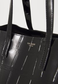 Saco tote preto brilhante com textura em relevo de crocodilo e logotipo dourado da Ted Baker London perto do topo, apresentando duas alças para os ombros.