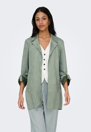 Žena u svijetlozelenom otvorenom sakou s podvrnutim rukavima preko bijele košulje s gumbima i svijetlosivim hlačama, stoji nasuprot običnoj sivoj pozadini.