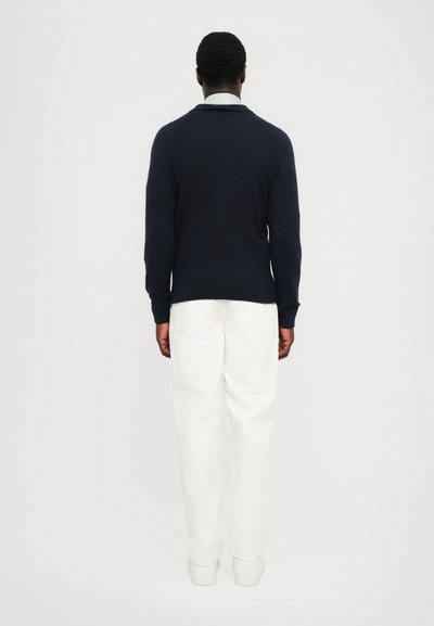 Cárdigan azul marino con dobladillo y cuello acanalados, fabricado con material suave, usado sobre pantalones blancos, visto desde la parte posterior.