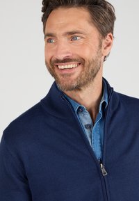 Marineblauer Zip-Pullover mit Stehkragen, gestrickter Textur und silbernem Reißverschluss, über einem hellblauen Hemd mit Knöpfen getragen.