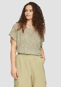 Olijfgroene top met witte stippenpatroon, losse pasvorm, korte mouwen en een brede halslijn, gecombineerd met lichtbeige broek met elastische tailleband.