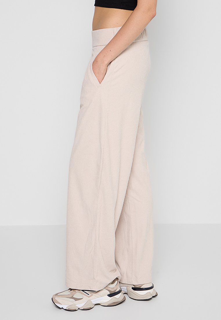 Donna che indossa pantaloni larghi beige e sneaker chunky beige, in piedi con una mano in tasca su uno sfondo semplice.