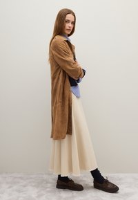 Cappotto lungo in suede beige sopra una camicia a righe blu e bianche, gonna maxi di colore crema e scarpe marroni con calze nere a coste. Texture lisce, look a strati.