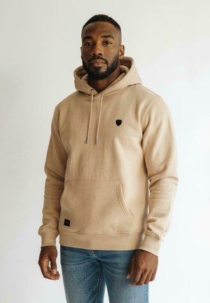 Homme avec une barbe portant un sweat à capuche beige et un jean bleu, se tenant devant un fond clair uni, faisant face à l'avant avec une expression neutre.