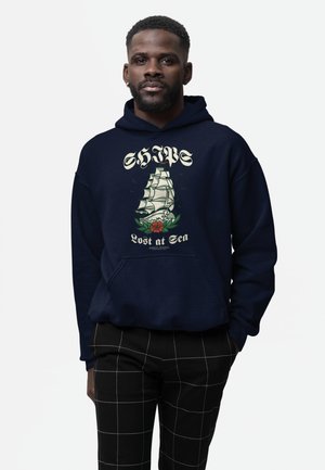 Man draagt een marineblauwe hoodie met een zeilschipillustratie en de tekst "SHIP Lost at Sea", gecombineerd met zwarte broek met wit ruitpatroon.