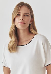 White top with a wide round neckline, short sleeves, and navy blue trim along the edges. Smooth fabric texture, casual design.