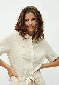 Vestido camisa de manga corta beige con cuello, dos bolsillos en el pecho, cintura ajustada y tejido texturizado sobre un fondo claro.
