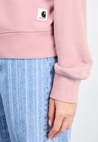 Sweatshirt rose clair avec des poignets et un ourlet côtelés, arborant une étiquette carrée blanche avec un logo noir. Associé à un jean à rayures verticales bleu clair.