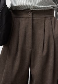 Pantaloni in corduroy marrone con pieghe, chiusura a un bottone in vita e tasche laterali. La texture del tessuto è a coste e morbida.