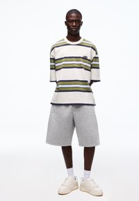 Unge mand stående iført en stribet kortærmet skjorte, grå knælange shorts, hvide sokker og beige sneakers på en hvid baggrund.