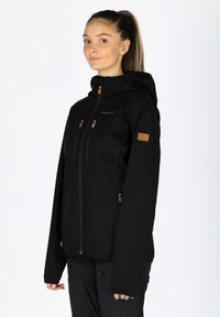 Svart mjukskaljacka med huva, med zippered fickor och bruna detaljer på bröstet och ärmen. Slät textur, designad för utomhusbruk.