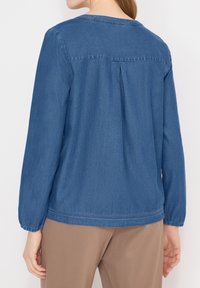 Femme portant une chemise en denim bleue à manches longues avec des poignets froncés et un pantalon beige, vue de dos sur un fond uni.