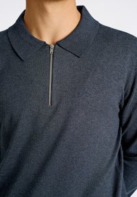 Tummansininen vetoketjullinen poolopaita, jossa on neulottu pinta, ja vasemmassa rinnassa pieni brodeerattu logo.