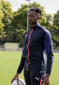 Puma trenirka v mornarsko modri barvi s vijoličnimi poudarki in roza zadrgo. Model drži nogometno žogo na travnati površini.