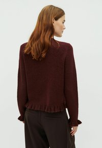 Burgundský pletený sveter s textúrovaným povrchom a volánovým lemom. Má dlhé rukávy s volánovými manžetami. Kombinuje sa s tmavými nohavicami.