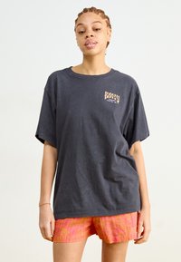 Overdimensjonert mørkegrå t-skjorte med en liten lys gul logo. Kombinert med teksturerte rosa og oransje rutete shorts. Mykt bomullsmateriale.