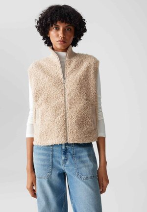 Donna con capelli ricci indossa un gilet beige texturizzato con cerniera sopra una maglia bianca a maniche lunghe e jeans blu a vita alta su uno sfondo grigio semplice.