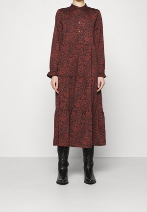 Femme portant une robe midi à manches longues de couleur rouge rouille avec un motif floral noir et des bottes en cuir noires arrivant au mollet, se tenant devant un fond uni.
