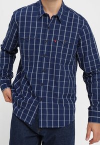 Chemise à carreaux bleu marine à manches longues, à boutonnage frontal, et deux poches poitrine. Comprend des boutons sombres et une étiquette logo discrète sur la poche.