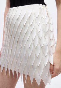 Mini-jupe blanche avec des pièces de tissu superposées en forme de pétales, portée avec un haut noir, montrant une fermeture éclair et une main reposant sur le côté.