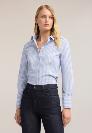 Kvindelig blond med en lyseblå stribet skjorte med knapper, stoppet ned i højttaljede mørke jeans, stående foran en neutral baggrund.