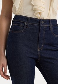 Jeans i denim med en mörkblå färg, hög midja, subtila orangea sömmar och framfickor. Tyget har en slät, strukturerad ytfinish.