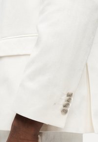 Σκούρο δέρμα σε χέρι που φοράει λευκό σακάκι με τέσσερα μπεζ κουμπιά στην μανσέτα και οριζόντια ραφή κοντά στη μέση.