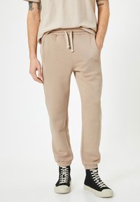 Mand iført beige sweatpants med snor og elastiske manchetter, parret med sorte højtop sneakers med hvide snørebånd på hvid baggrund.