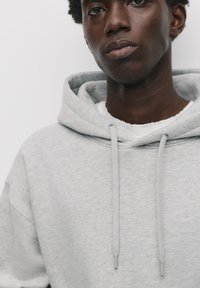 Felpa grigia chiara con cappuccio e coulisse, realizzata in un tessuto morbido e texturizzato e con una vestibilità comoda. Indossata sopra una maglietta bianca a girocollo.