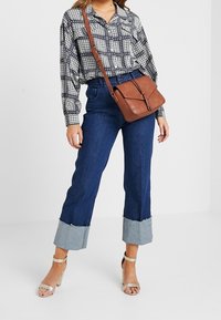 Brązowa skórzana torebka crossbody z plecionym akcentem, zestawiona z czarno-białą koszulą w wzory oraz skróconymi niebieskimi dżinsami z postrzępionymi mankietami.