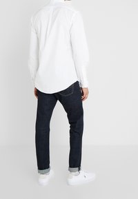 Biała koszula z długim rękawem o zaokrąglonym krawędzi, w parze z ciemnoniebieskimi jeansami. Model nosi białe trampki i podwinięte mankiety.